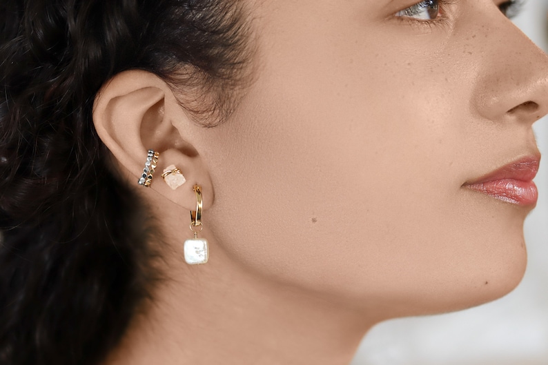 K&ouml;nnte beinhalten: Nahaufnahme eines Ohrs, das einen goldenen Creolenohrring mit einem wei&szlig;en Perlenanh&auml;nger, einen goldenen Ohrclip mit einem kleinen rosafarbenen Stein und einen silbernen und goldenen Ohrclip tr&auml;gt.