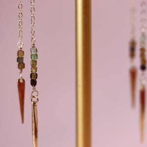 May include: A pair of gold earrings with a long, thin, pointed pendant and a chain of green, brown, and black beads.