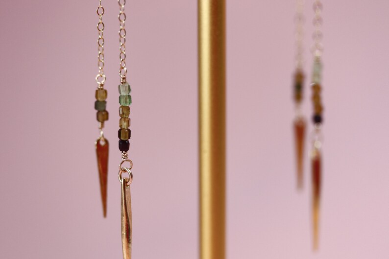 May include: A pair of gold earrings with a long, thin, pointed pendant and a chain of green, brown, and black beads.