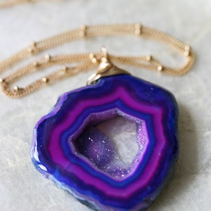 Peut inclure: Un pendentif en agate violette avec une chaîne dorée. L'agate a un trou naturel au centre et est entourée d'une bande rose et bleue.