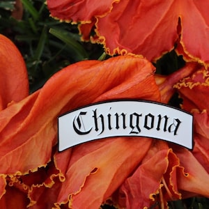May include: White enamel pin with black lettering that reads "Chingona" in a gothic font. The pin is shaped like a banner and is sitting on a bright orange flower.