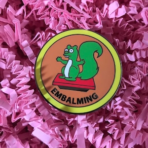 May include: A yellow and orange enamel pin featuring a green cartoon squirrel standing on a red platform with the word "EMBALMING" written in black letters.