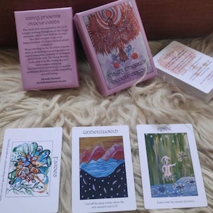 Puede incluir: Un juego de "Rising Phoenix Oracle Cards" en una caja rosa, con tres cartas expuestas. Las cartas presentan ilustraciones coloridas y las palabras "Fairies", "Underworld" y "Spirit". La caja tiene texto y una ilustración de fénix.