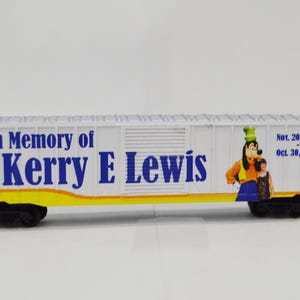 May include: A white model train car with blue lettering that reads "In Memory of Kerry E Lewis" and a cartoon image of Goofy and a child. The dates "Nov. 20, 1948" and "Oct. 30, 2019" are also printed on the car.