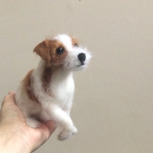 May include: A white and brown needle felted Jack Russell Terrier dog figurine. The dog is sitting with its head tilted up and its eyes looking forward.