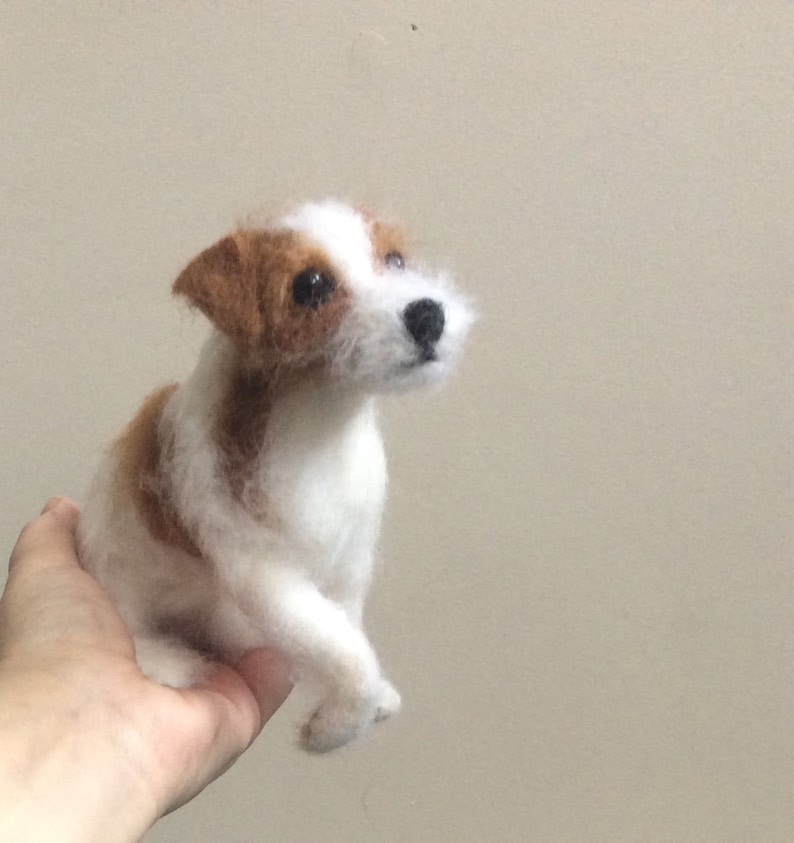 May include: A white and brown needle felted Jack Russell Terrier dog figurine. The dog is sitting with its head tilted up and its eyes looking forward.