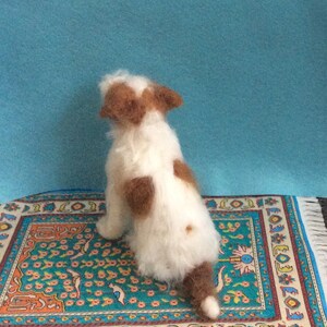 May include: A white and brown needle felted dog figurine sitting on a colorful rug with a blue background.