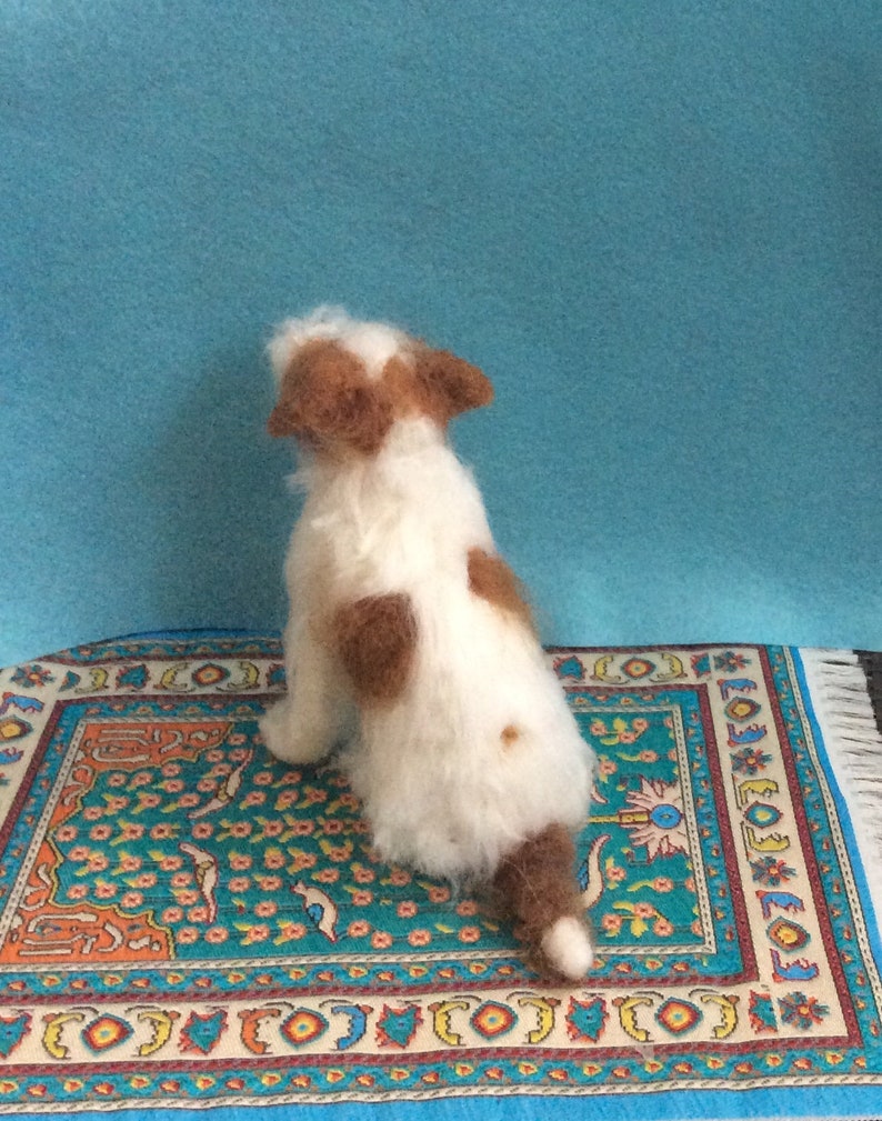 May include: A white and brown needle felted dog figurine sitting on a colorful rug with a blue background.