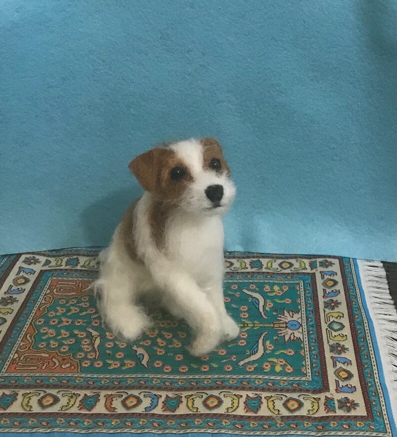 May include: A white and brown needle felted Jack Russell Terrier dog sitting on a colorful patterned rug.
