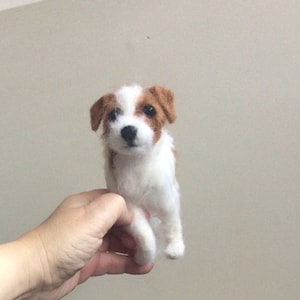 May include: A white and brown needle felted Jack Russell Terrier dog figurine. The dog is sitting up with its front paws crossed and looking directly at the camera.