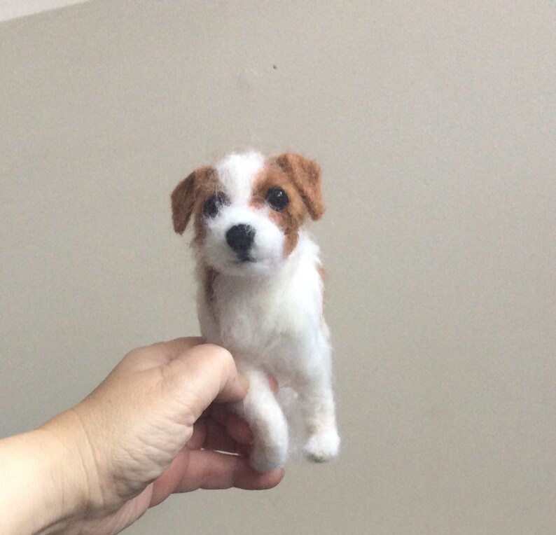 May include: A white and brown needle felted Jack Russell Terrier dog figurine. The dog is sitting up with its front paws crossed and looking directly at the camera.