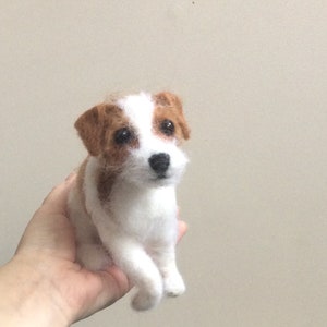 May include: A white and brown needle felted Jack Russell Terrier dog figurine. The dog is sitting with its front paws crossed and its head tilted slightly to the side.