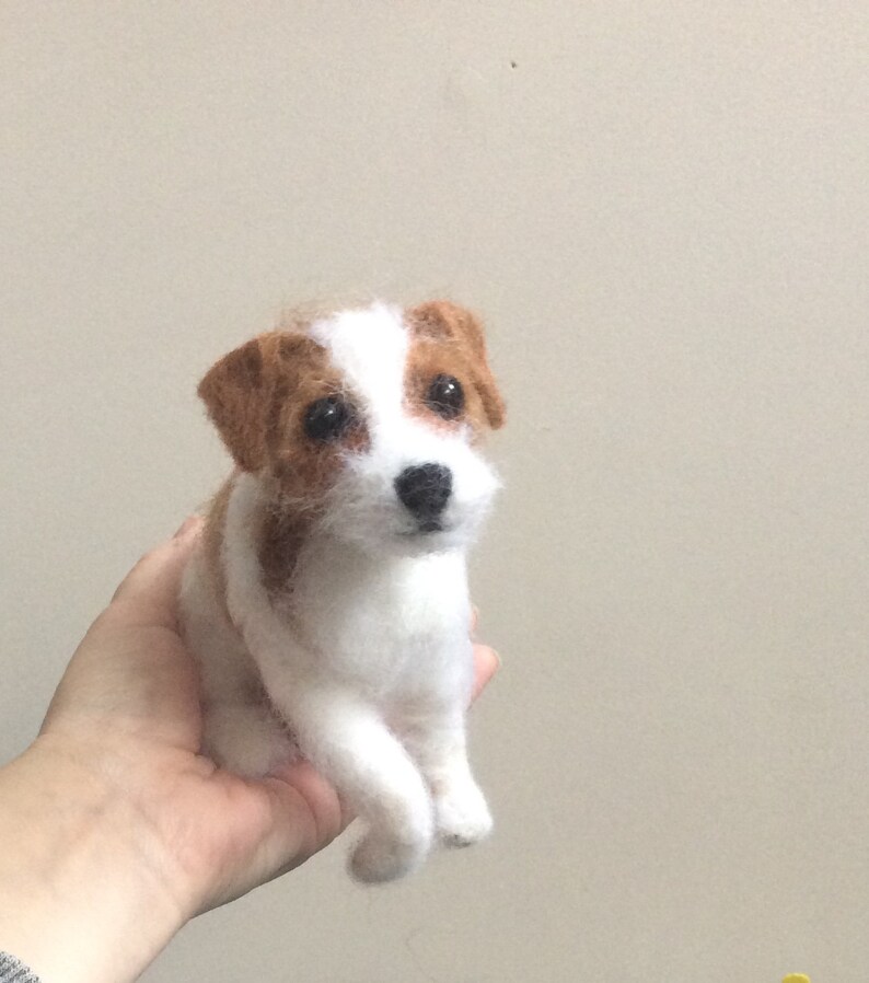 May include: A white and brown needle felted Jack Russell Terrier dog figurine. The dog is sitting with its front paws crossed and its head tilted slightly to the side.