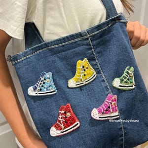 May include: A denim tote bag adorned with five colorful, beaded sneaker brooches. The brooches are in shades of blue, yellow, red, pink, and green, each with white soles and laces. The brooches are embellished with beads and sequins.