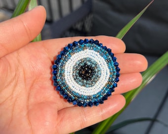 May include: A round, beaded amulet featuring a central eye design. The eye is composed of concentric circles in white, light blue, dark blue, and black, surrounded by a border of dark blue beads. The amulet is held in a hand.