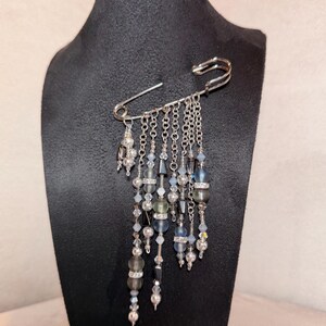 May include: A large silver safety pin with dangling beaded chains. The chains feature beads in blue, grey, and clear, with silver accents. The pin is displayed on a black mannequin.