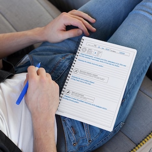 May include: A white spiral-bound notebook with a detailed checklist, held by a person wearing blue jeans. The checklist includes sections for annual maintenance, spark plugs, and brake checks. A blue pen is held near the notebook.