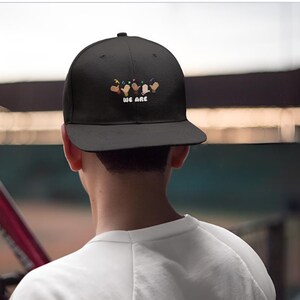 May include: Black baseball cap with a flat brim. The cap features an embroidered design of hands in various skin tones, with colorful confetti and the text "WE ARE" in white.