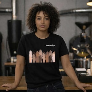 May include: Black t-shirt with the word "Humanity" printed in white. The shirt features a graphic of hands in various skin tones raised in the air. The model is wearing the shirt and is standing in front of a wooden table.