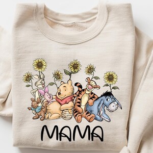 Puede incluir: Una sudadera de color crema con un estampado gr&aacute;fico de Winnie de Pooh y sus amigos, incluyendo Tigger, Piglet y Eeyore, rodeados de girasoles. El texto "MAMA" est&aacute; impreso debajo del gr&aacute;fico.