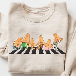 May include: Cream-colored sweatshirt featuring a playful graphic of cartoon characters crossing a black and white striped crosswalk, each wearing an orange traffic cone. The long-sleeved top is folded on a white surface.