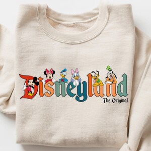 Może przedstawiać: Beżowa bluza z logo Disneyland z Myszka Miki, Myszka Minnie, Kaczorem Donaldem, Daisy, Goofym i Plutonem. Tekst "The Original" znajduje się pod tekstem Disneyland. Bluza jest złożona.