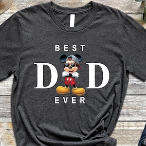 Op de afbeelding: Een donkergrijs T-shirt met een witte afbeelding van Mickey Mouse met een zonnebril en een petje achterstevoren. De tekst "BEST DAD EVER" is in wit onder de afbeelding gedrukt.