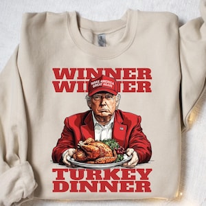 May include: A beige sweatshirt with a red graphic print of a man wearing a red "Make America Great Again" hat holding a plate of roasted turkey. The text on the sweatshirt reads "Winner Winner Turkey Dinner".
