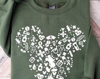 May include: An olive green sweatshirt featuring a white Mickey Mouse head design filled with medical-themed icons. The design includes syringes, pills, and other healthcare symbols. The sweatshirt has cuffed sleeves and a crew neck.
