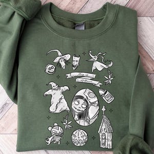 K&ouml;nnte beinhalten: Ein olivgr&uuml;nes Sweatshirt mit einem schwarz-wei&szlig;en Design mit Figuren und Elementen aus einem Halloween-Film. Das Design umfasst Charaktere, ein Banner mit Text und einen K&uuml;rbis.