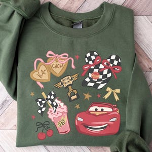 May include: Green sweatshirt featuring a colorful graphic design. The design includes a red race car, checkered flags, a milkshake, and the text "KA-CHOW". The sweatshirt has cuffed sleeves and a crew neck.