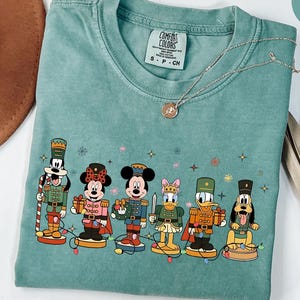 May include: Light green t-shirt featuring a line of cartoon characters dressed as nutcrackers. The characters include Goofy, Minnie Mouse, Mickey Mouse, Daisy Duck, Donald Duck, and Pluto. The shirt has a Comfort Colors tag.