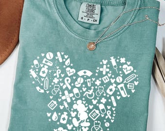 May include: Light green t-shirt with a white Mickey Mouse head design filled with medical symbols. The shirt has a crew neck and short sleeves. A gold necklace with a small pendant is draped across the shirt.