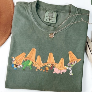 Puede incluir: Camiseta verde musgo con un gr&aacute;fico de personajes de dibujos animados que llevan conos de tr&aacute;fico naranjas. La camiseta tiene cuello redondo y mangas cortas. Un collar dorado con un colgante est&aacute; sobre la camiseta.