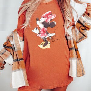 Puede incluir: Una camiseta de color &oacute;xido con un gr&aacute;fico de Minnie Mouse con un vestido rojo de lunares y un lazo rojo. El texto "Minnie Mouse" est&aacute; impreso en marr&oacute;n debajo del gr&aacute;fico.