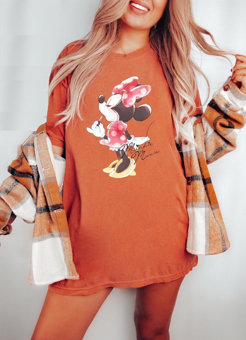 Puede incluir: Una camiseta de color &oacute;xido con un gr&aacute;fico de Minnie Mouse con un vestido rojo de lunares y un lazo rojo. El texto "Minnie Mouse" est&aacute; impreso en marr&oacute;n debajo del gr&aacute;fico.