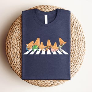 May include: Navy blue t-shirt featuring a cartoon illustration of several characters wearing orange traffic cones and crossing a white and black crosswalk. The characters are in various colors and designs. The shirt is folded on a woven surface.