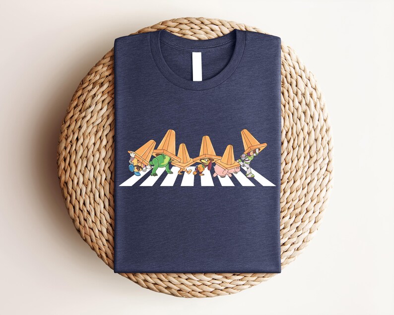 May include: Navy blue t-shirt featuring a cartoon illustration of several characters wearing orange traffic cones and crossing a white and black crosswalk. The characters are in various colors and designs. The shirt is folded on a woven surface.