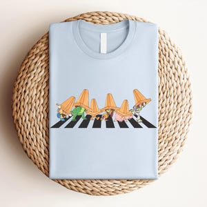 May include: Light blue t-shirt featuring a graphic of cartoon characters crossing a black and white crosswalk, each wearing an orange traffic cone on their head. The design is centered on the shirt.