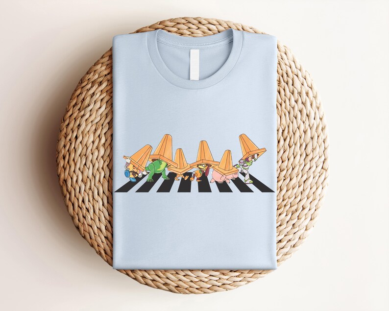 May include: Light blue t-shirt featuring a graphic of cartoon characters crossing a black and white crosswalk, each wearing an orange traffic cone on their head. The design is centered on the shirt.