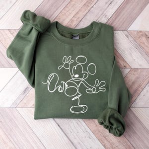 Puede incluir: Una sudadera verde con un contorno blanco de Mickey Mouse haciendo malabares. El dise&ntilde;o es simple y minimalista.