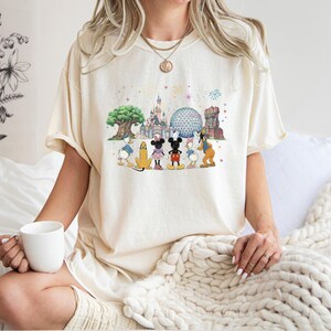 May include: Cream-colored t-shirt featuring a colorful graphic of Disney characters and park icons. The design includes Mickey Mouse, Minnie Mouse, Donald Duck, and Goofy, with Cinderella's castle and Spaceship Earth in the background. The shirt is worn by a person.