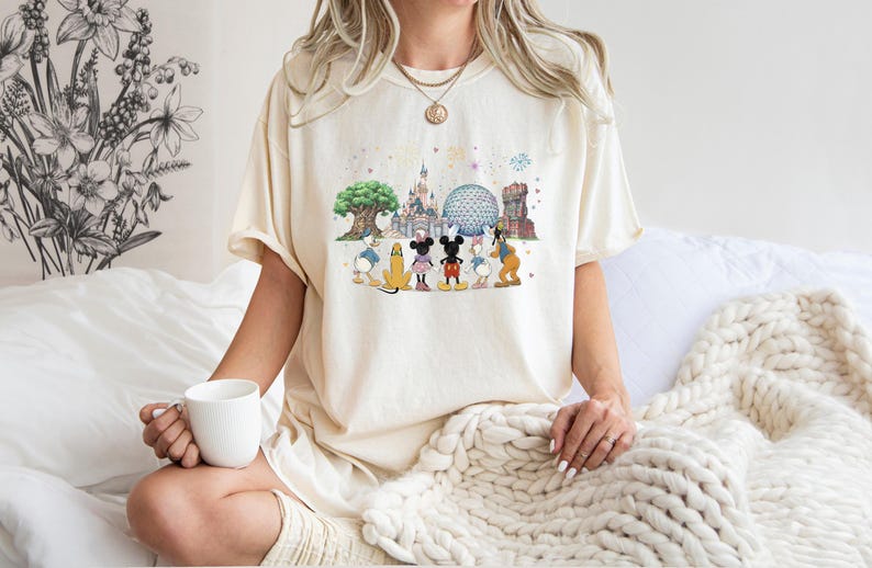 May include: Cream-colored t-shirt featuring a colorful graphic of Disney characters and park icons. The design includes Mickey Mouse, Minnie Mouse, Donald Duck, and Goofy, with Cinderella's castle and Spaceship Earth in the background. The shirt is worn by a person.