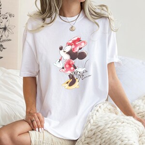 Puede incluir: Camiseta blanca de gran tama&ntilde;o con un estampado gr&aacute;fico de Minnie Mouse con un vestido rojo con lunares blancos y un lazo rojo. El texto "Minnie Mouse" est&aacute; impreso debajo del gr&aacute;fico.