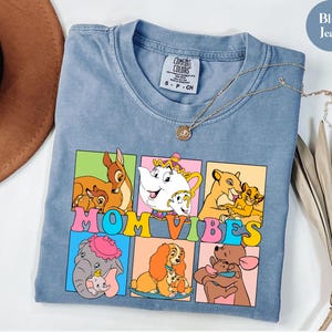 May include: Blue jean t-shirt with a graphic of Disney characters in colorful squares. The text "MOM VIBES" is printed in rainbow colors across the squares.