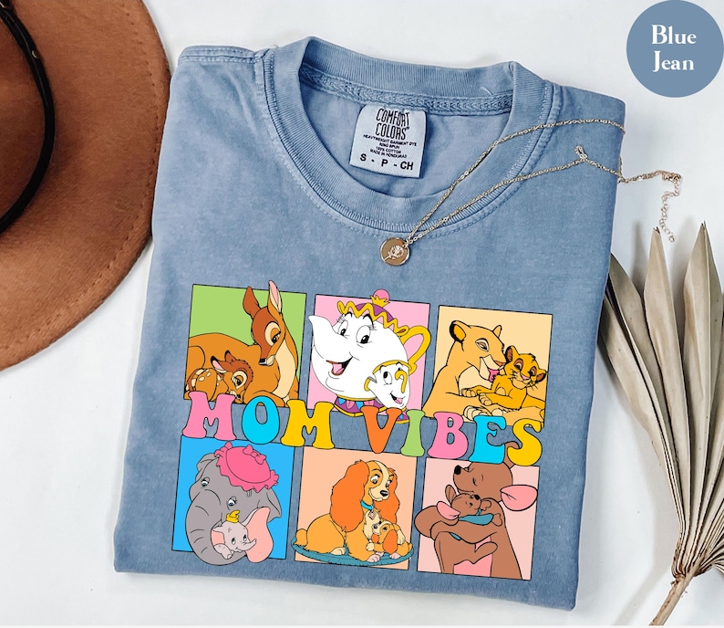 May include: Blue jean t-shirt with a graphic of Disney characters in colorful squares. The text "MOM VIBES" is printed in rainbow colors across the squares.