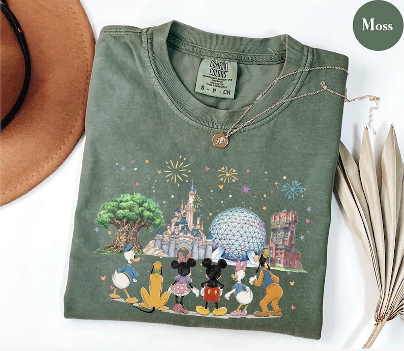 May include: Moss green t-shirt featuring a colorful graphic of Disney characters and landmarks, including Cinderella's castle and Spaceship Earth. The shirt has a crew neck and short sleeves. A gold necklace is draped across the shirt.