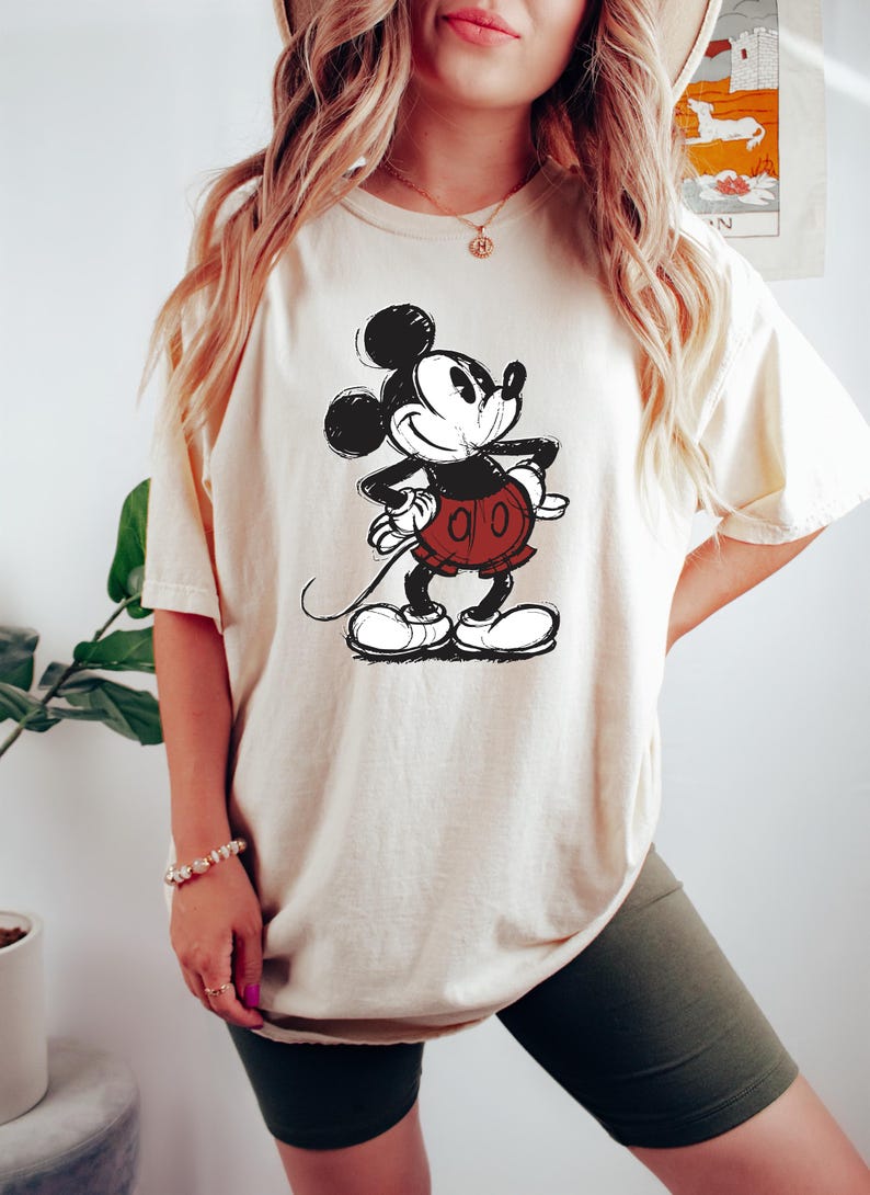May include: A cream colored t-shirt with a black and white sketch of Mickey Mouse wearing red shorts. The t-shirt is oversized and has a relaxed fit.