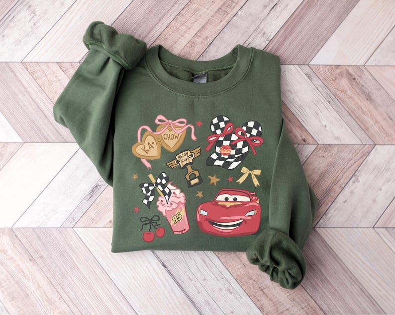 May include: Green sweatshirt featuring a colorful graphic design. The design includes a red race car, checkered flags, a milkshake, and the text "KA-CHOW". The sweatshirt has cuffed sleeves and a crew neck.