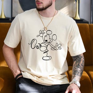 Puede incluir: Una camiseta blanca con un dibujo de l&iacute;nea negra de Mickey Mouse haciendo malabares con c&iacute;rculos.